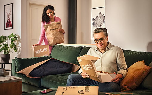 man sitting on sofa smiling while opening a package as a lady stands behind holding packaging